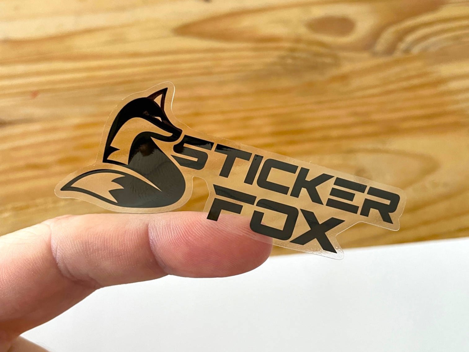 StickerFox logo sticker held on a fingertip against a wooden background